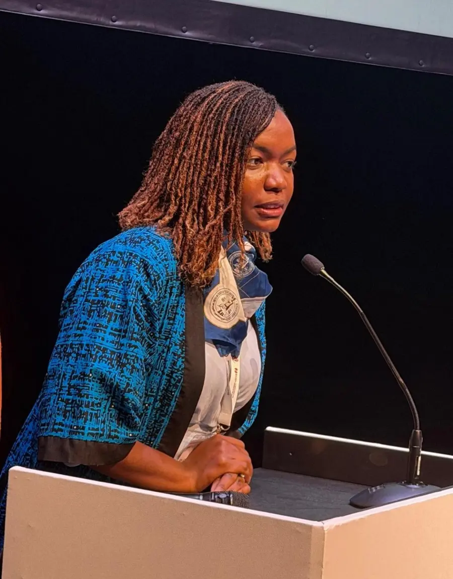 Dr Christine Nyabuto speaking at a lectern microphone, wearing a blue patterned jacket and a conference lanyard.